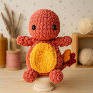Könnte beinhalten: Ein handgefertigtes, gehäkeltes Plüschtier in Form eines Zeichentrickdrachen. Das Spielzeug ist hauptsächlich korallenfarben mit einem gelben Bauch und einem Schwanz mit orangefarbenen und roten Akzenten. Es hat schwarze Sicherheitsaugen und sitzt auf einem kleinen Holzsockel.