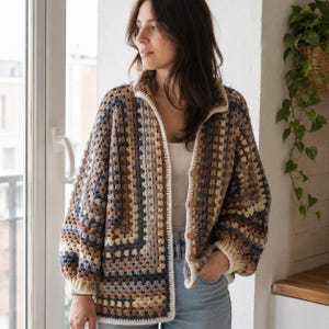 Puede incluir: Chaqueta de ganchillo multicolor con un patrón de cuadrados de la abuela. La chaqueta presenta tonos de marrón, beige y azul, con cuello y ribete color crema. Tiene mangas largas y botones de madera en la parte delantera, ofreciendo una estética artesanal y acogedora.