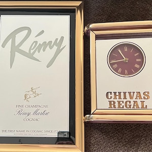 May include: Two gold-framed wall decorations. One displays the Rémy logo and text, the other a clock with the Chivas Regal logo. The clock has a burgundy face and gold hands.