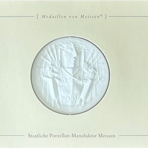 Peut inclure: Médaillon en porcelaine blanche de Meissen, représentant en relief une figure classique aux bras tendus, daté de 2001. Le médaillon est encadré dans une carte crème avec du texte en haut et en bas.