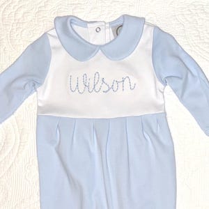 May include: A light blue baby romper with long sleeves and footed bottoms. The romper features a white bib-like panel with the name "Wilson" embroidered in blue. It has a rounded collar and button closure.