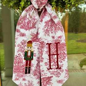 May include: White and red toile Christmas table runner with a nutcracker and the letter "H" embroidered on it. The runner is hanging from a green wreath. The toile pattern depicts scenes of people and buildings.