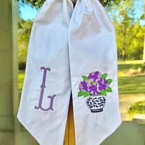 May include: White fabric sash with a purple embroidered initial "L" on one side and a vase of purple flowers on the other. The vase has a blue and white floral design. The sash is hanging in front of a green wreath.