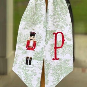 May include: A white fabric hanging decoration featuring a green toile pattern of trees and figures. One side displays an embroidered nutcracker in red, black, and white, while the other side features a large, embroidered red letter P in serif font.
