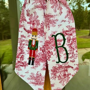 May include: White fabric Christmas decoration with a red toile pattern and a green embroidered letter "B". The fabric features a nutcracker and a decorative design. The decoration is hanging on a window.