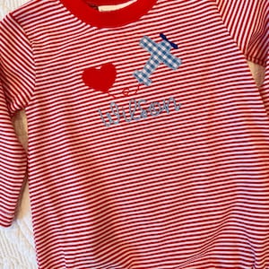 May include: Red and white striped long-sleeved onesie with a red collar. Features a blue gingham airplane applique, a red heart, and the name "Wilson" embroidered in blue script. A charming outfit for a baby or toddler.
