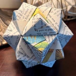 Pode incluir: Uma escultura geométrica de origami feita com páginas de livro. O papel é dobrado em forma de estrela, com texto e ilustrações visíveis. Está sobre uma superfície de madeira escura.