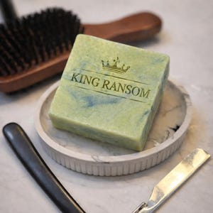 Op de afbeelding: Een groen en blauw zeepblok met de tekst "KING RANSOM" en een kroonlogo, rustend op een marmeren dienblad. Een recht scheermes en een houten borstel zijn ook zichtbaar.