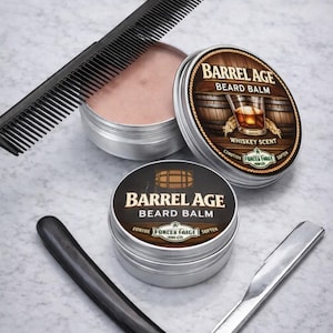 以下が含まれることがあります： 「Barrel Age」のひげバームの銀色の缶2つ、黒い櫛、ストレートカミソリなど、グルーミング製品の詰め合わせ。バーム缶には、ウイスキーグラスのイラストと「Whiskey Scent」の文字が入った茶色のラベルが付いています。