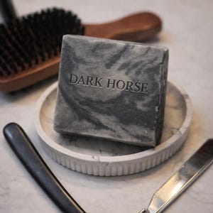 Könnte beinhalten: Ein dunkelgraues Seifenstück mit dem geprägten Text "DARK HORSE" liegt auf einem Marmortablett. Eine Holzbürste und ein Rasiermesser sind ebenfalls zu sehen. Die Seife hat ein marmoriertes Muster.