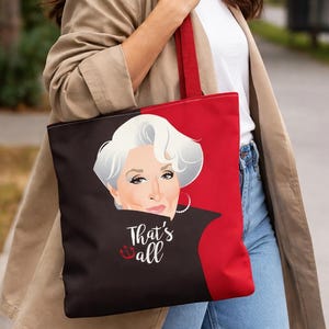 Pode incluir: Bolsa tote vermelha e preta com um retrato de uma mulher com cabelo branco. O texto "That's all" está escrito em branco com um símbolo de âncora. A bolsa tem alças vermelhas.