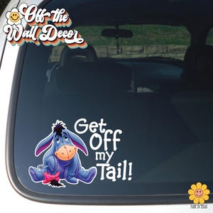 May include: A car window decal featuring Eeyore from Winnie the Pooh, in shades of blue, pink, and tan. The decal reads "Get Off my Tail!" in white text. The decal is from "Off the Wall Decor."