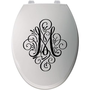 May include: White toilet seat with a black, ornate letter "M" design. The letter is stylized with swirling lines and decorative flourishes. The seat has a standard oval shape and is made of a smooth, glossy material.