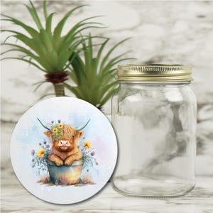 May include: A clear glass jar with a gold lid next to a round coaster. The coaster features a watercolor illustration of a Highland cow in a flower pot, adorned with a floral crown and surrounded by yellow and purple flowers.