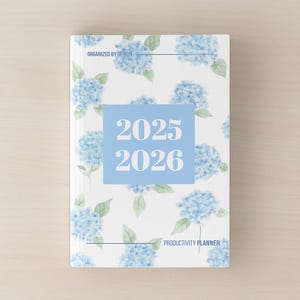 May include: A hardcover planner with a white background and a pattern of blue hydrangea flowers and green leaves. The planner cover has a light blue square with the numbers "2025 2026" in white. The text "Organized by Design" and "Productivity Planner" are also visible.