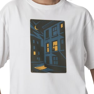 May include: White t-shirt featuring a graphic illustration of a building at night. The artwork depicts a building with lit windows, a crescent moon, and a dark blue and yellow color scheme. The shirt is made of a soft, comfortable material.