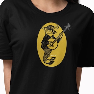 May include: Black t-shirt featuring a cartoon character playing a banjo inside a yellow oval. The character is a frog wearing a jacket and hat. The design is in black and white. The person wearing the shirt is wearing white headphones.