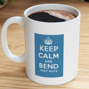 May include: White ceramic coffee mug with a blue rectangle featuring a crown graphic and the text "KEEP CALM AND BEND THAT NOTE". The mug is filled with dark coffee and sits on a wooden surface.