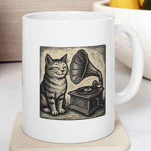 May include: White ceramic mug featuring a black and white illustration of a smiling cat next to a vintage gramophone. The mug has a curved handle and is sitting on a beige coaster. The design has a vintage, whimsical feel.