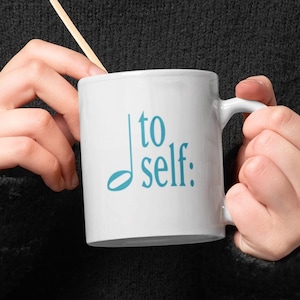 May include: White ceramic mug with the text "to self:" in teal. A wooden stirrer rests inside the mug. The mug is held by a person wearing a black sweater. The mug is a standard 11 oz size.