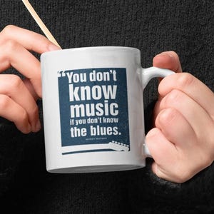 May include: White ceramic mug with the text "You don't know music if you don't know the blues." The mug has a handle and a wooden stirrer. The mug is held by a person wearing a black sweater.