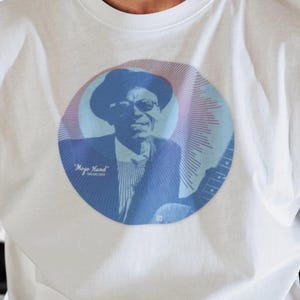 Puede incluir: Camiseta blanca con un gráfico circular de un hombre con sombrero y traje, con el texto "Mojo Hand". La persona de la imagen lleva un sombrero fedora beige y gafas redondas.