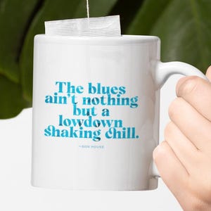 May include: White ceramic mug with the quote "The blues ain't nothing but a lowdown shaking chill" in blue text. A tea bag is suspended above the mug. The background features large green leaves.