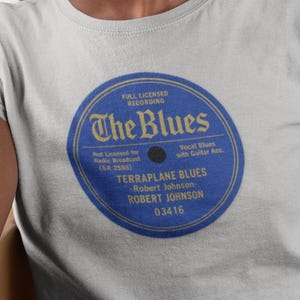 Könnte beinhalten: Hellgraues T-Shirt mit einem blau-goldenen Grafikdesign eines Vintage-Plattenlabels. Das Label trägt die Aufschrift "The Blues" mit Text wie "Terrplane Blues" und "Robert Johnson". Das T-Shirt wird von einer Person getragen.