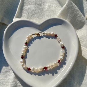 May include: A pearl bracelet with red and gold accents rests on a white heart-shaped plate. The bracelet features irregularly shaped white pearls interspersed with small red beads and gold-colored spacers. The clasp is gold-toned.