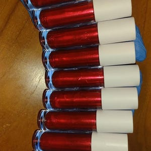 Könnte beinhalten: Eine Ansammlung von Lipgloss-Tuben mit leuchtend roter Farbe und weißen Kappen. Der Text "Ruby Rush" ist oben zu sehen. Die Tuben sind in einem Stapel angeordnet, was auf eine Kosmetikpräsentation hindeutet.