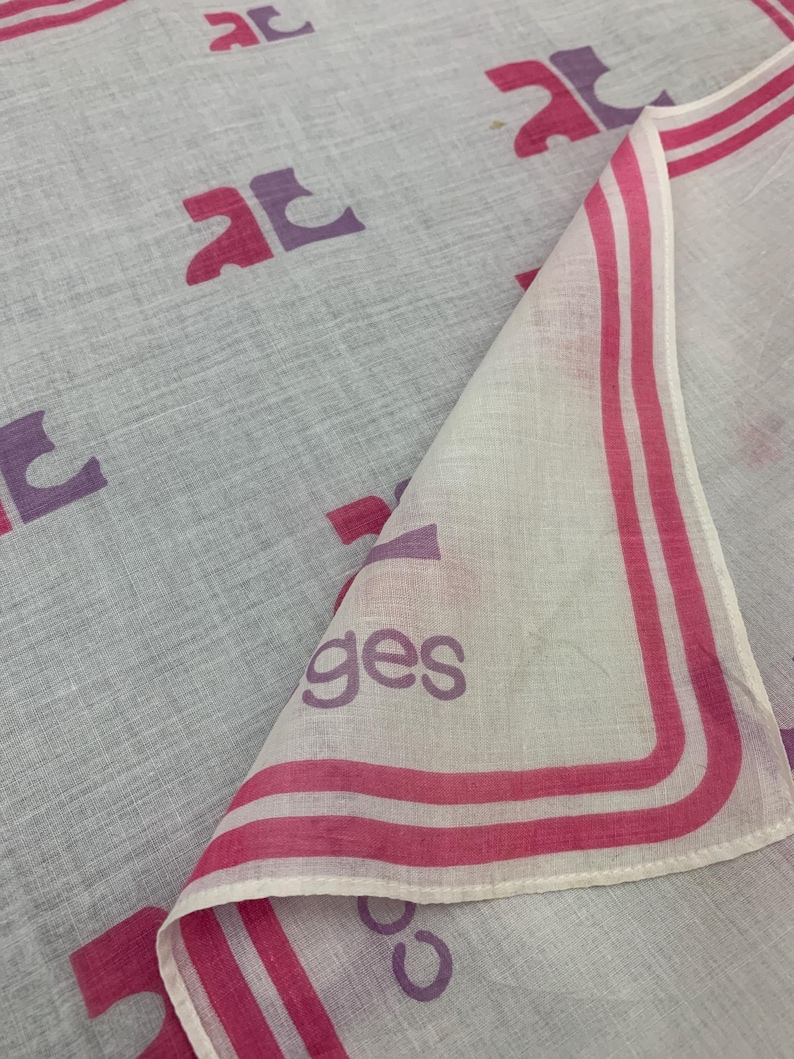 Peut inclure: Un foulard gris clair et blanc avec un motif r&eacute;p&eacute;t&eacute; de lettres stylis&eacute;es roses et violettes. Le bord du foulard a une bordure rose avec le mot "ges" imprim&eacute; en violet.