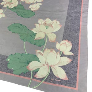 May include: A square, light purple scarf with a floral design. The scarf features large, white lotus flowers with pink edges and green leaves. A darker purple border with a pink trim surrounds the floral pattern. The scarf is likely made of a lightweight fabric.