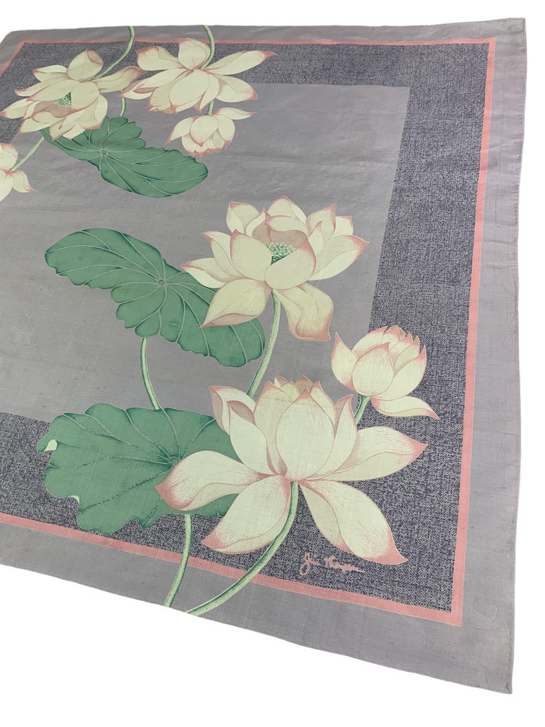May include: A square, light purple scarf with a floral design. The scarf features large, white lotus flowers with pink edges and green leaves. A darker purple border with a pink trim surrounds the floral pattern. The scarf is likely made of a lightweight fabric.