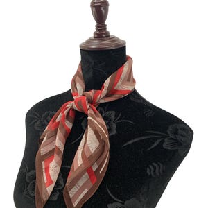 May include: A patterned scarf with a geometric design in shades of red, brown, and beige is draped around a mannequin's neck. The scarf is knotted at the front, showcasing its intricate pattern and color palette. The mannequin is black.