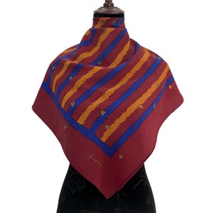 May include: A square scarf with a burgundy border and a pattern of orange and blue diagonal stripes. The scarf has small gold-colored accents and the Givenchy logo in cursive script.