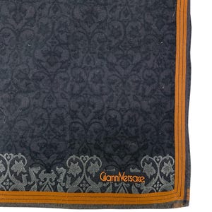 Puede incluir: Tela gris oscuro estampada con un borde naranja. La tela presenta un dise&ntilde;o floral repetitivo. Las palabras "Gianni Versace" est&aacute;n impresas en naranja. La tela est&aacute; doblada, mostrando una esquina del art&iacute;culo.
