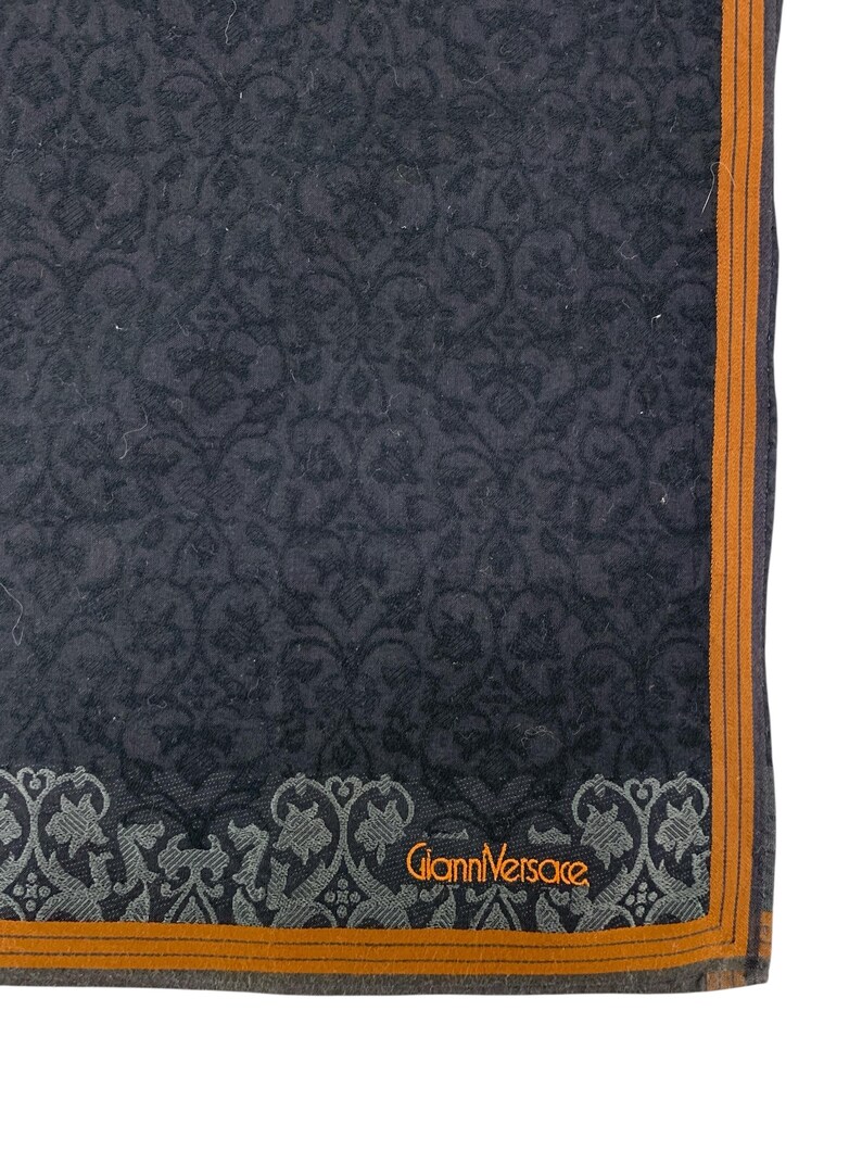 Puede incluir: Tela gris oscuro estampada con un borde naranja. La tela presenta un dise&ntilde;o floral repetitivo. Las palabras "Gianni Versace" est&aacute;n impresas en naranja. La tela est&aacute; doblada, mostrando una esquina del art&iacute;culo.