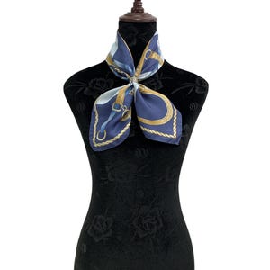 May include: A navy blue silk scarf with a gold and white equestrian design. The scarf is draped around a black mannequin. The scarf features a repeating pattern of gold rope, buckles, and straps.