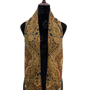 May include: A paisley patterned scarf in shades of gold, navy blue, and burgundy, displayed on a mannequin. The scarf features an intricate design with a fringed edge. The scarf is a rectangular shape and appears to be made of a soft material.