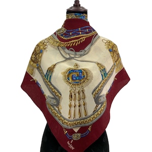May include: A square silk scarf with a cream-colored center and a deep red border. The scarf features a detailed design with gold, blue, and silver accents, including ornate patterns and a central emblem with dangling gold elements. The scarf is displayed on a mannequin.