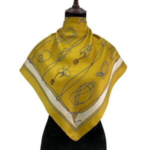 May include: Mustard yellow silk scarf with a rope and knot design. The scarf has a white border with a thin black outline. The scarf is displayed on a black mannequin. The scarf is square-shaped.