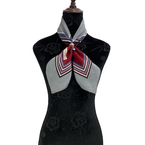 Könnte beinhalten: Ein graues, rotes und blaues Seidentuch mit geometrischem Muster, präsentiert auf einer schwarzen Schaufensterpuppe. Das Tuch ist geknotet und trägt den Schriftzug "YVES SAINT LAURENT".