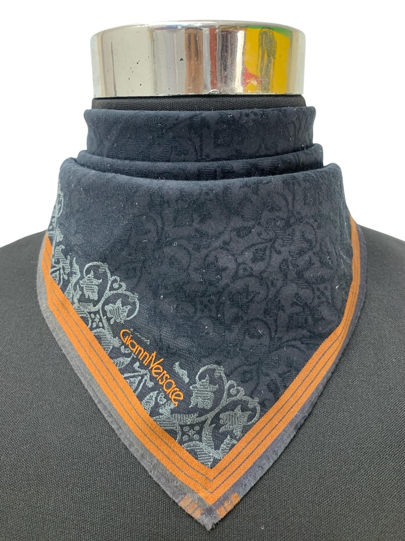 Puede incluir: Un pa&ntilde;uelo azul marino oscuro con un estampado floral adornado y un borde naranja. El pa&ntilde;uelo presenta el texto "Gianni Versace" en fuente plateada. El pa&ntilde;uelo se exhibe en un maniqu&iacute;.