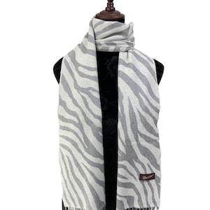 Peut inclure: Une longue écharpe présentant un motif à rayures zébrées gris clair et blanc. L'écharpe est de forme rectangulaire avec des extrémités à franges et une petite étiquette rectangulaire marron avec du texte doré cousue sur le côté droit.