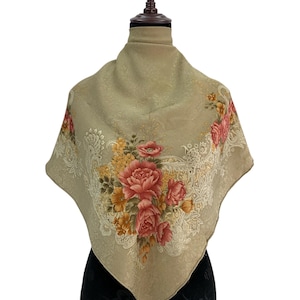 May include: A square, beige scarf featuring a floral pattern of pink and yellow roses. The scarf is displayed on a mannequin. The floral design is accented with white lace-like details. The scarf has a soft, flowing appearance.
