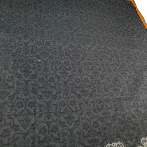 Puede incluir: Un textil gris oscuro con un patr&oacute;n floral repetitivo. El borde presenta un dise&ntilde;o decorativo en gris y blanco, con un ribete naranja. La tela tiene un aspecto suave y texturizado.