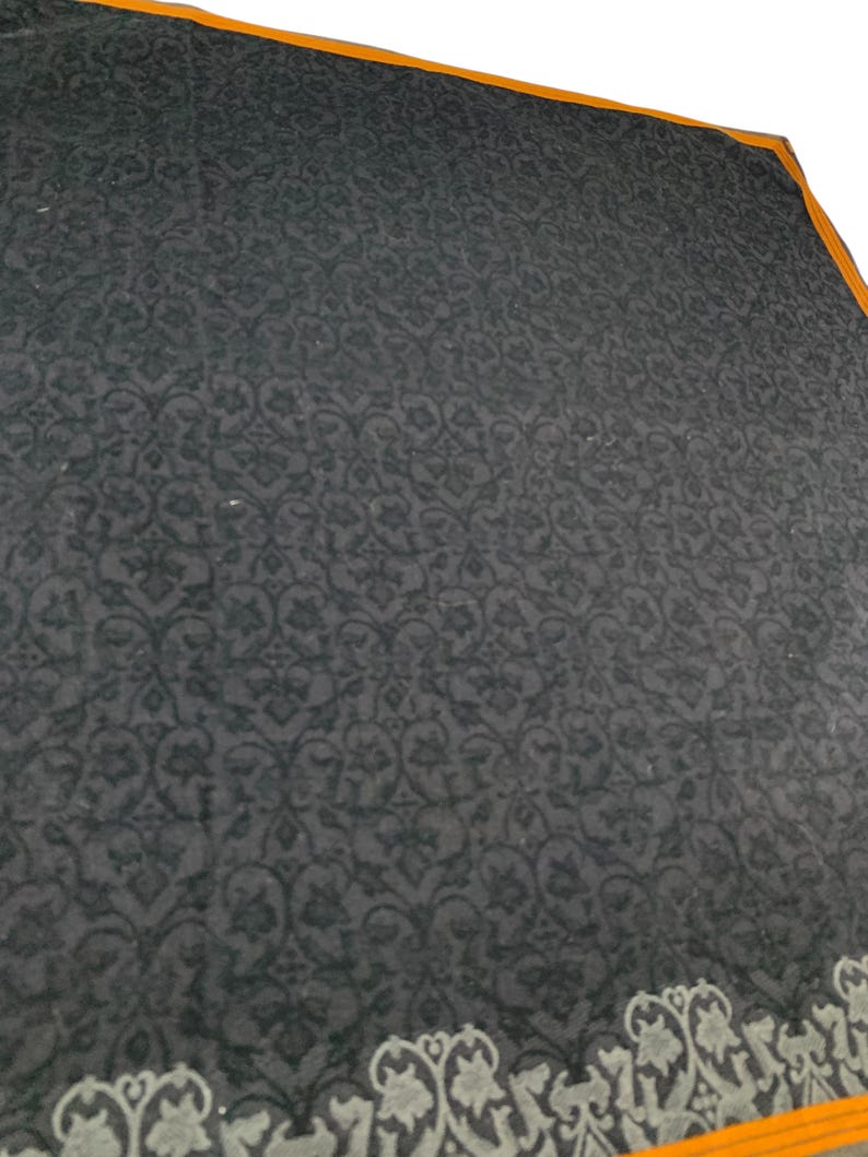 Puede incluir: Un textil gris oscuro con un patr&oacute;n floral repetitivo. El borde presenta un dise&ntilde;o decorativo en gris y blanco, con un ribete naranja. La tela tiene un aspecto suave y texturizado.