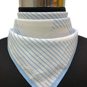 K&ouml;nnte beinhalten: Wei&szlig;-blau gestreifter Schal mit hellblauem Rand. Der Schal ist gefaltet und &uuml;ber einer schwarzen Oberfl&auml;che drapiert. Der Markenname "courr&egrave;ges" ist auf dem Schal zu sehen.