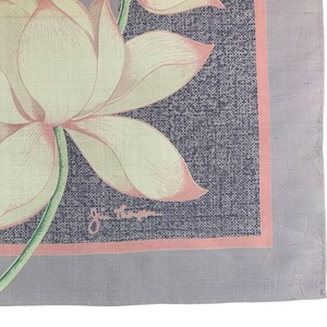 May include: A square, floral-patterned scarf featuring a large lotus flower with cream-colored petals and pink edges. The flower is set against a textured, dark blue background with a pink border, and a gray border. The scarf has a signature in the bottom right corner.