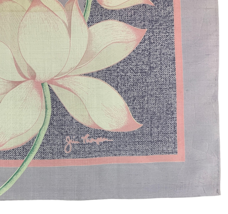 May include: A square, floral-patterned scarf featuring a large lotus flower with cream-colored petals and pink edges. The flower is set against a textured, dark blue background with a pink border, and a gray border. The scarf has a signature in the bottom right corner.
